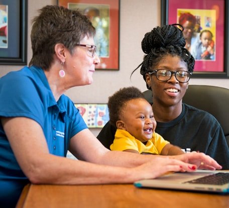 Goodwill volunteer giving computer training to a woman who is holding her smiling baby