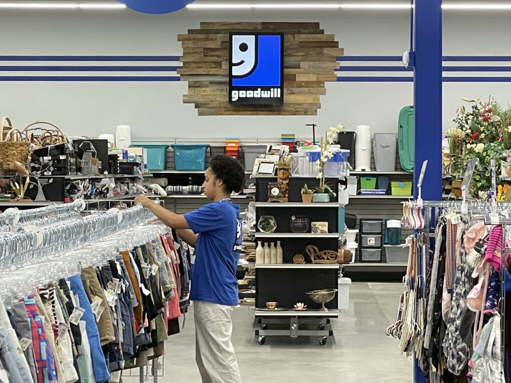 Goodwill employee next to rack of clothes with Smiling G logo against reclaimed wood on wall